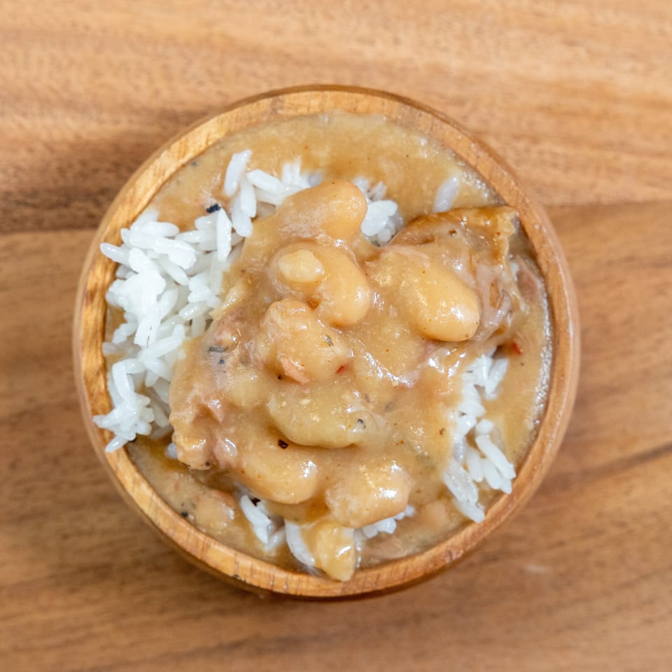Top-down view of white rice topped with creamy bean stew in a rustic wooden bowl on a wooden table.