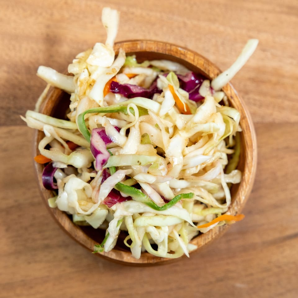 Bowl of shredded cabbage salad with purple cabbage, carrots, and green peppers on a wooden table.