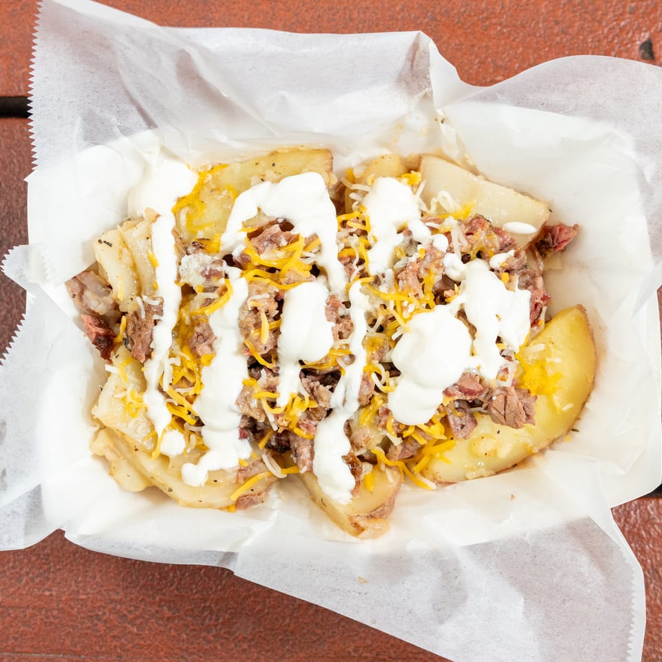 Loaded baked potato topped with chopped meat, shredded cheddar cheese, and creamy white sauce, served on parchment paper.