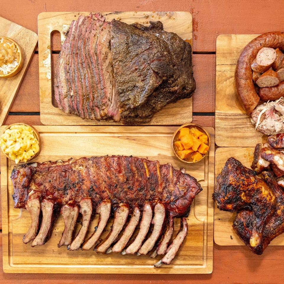 Overhead spread of smoked brisket, ribs, sausages, pulled pork, chicken, and barbecue sides arranged on wooden serving boards.