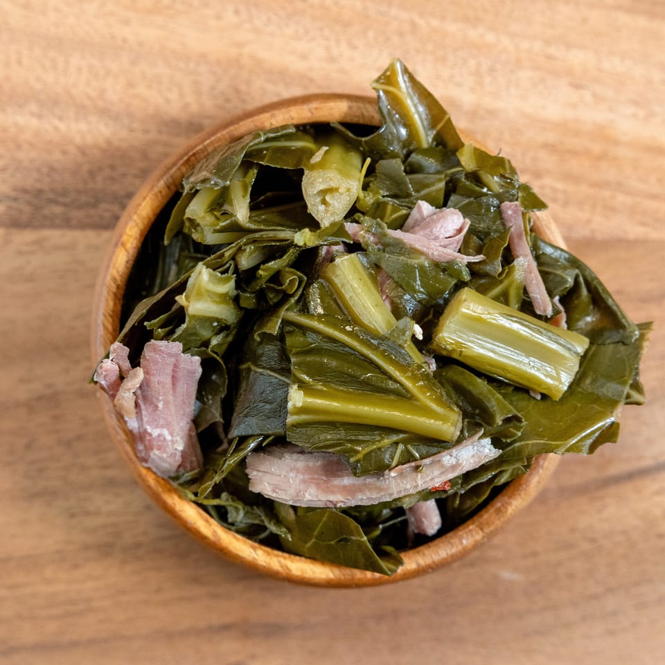 Bowl of cooked leafy greens with sliced ham on a wooden table.
