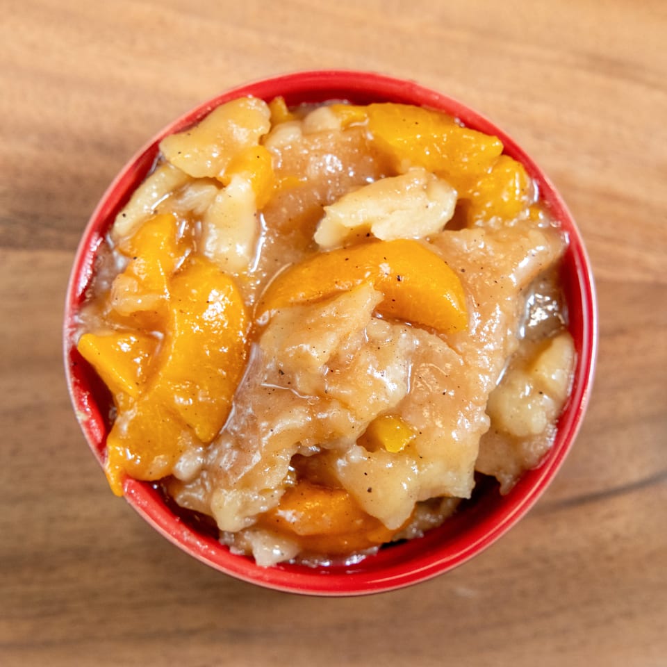 Top-down view of peach cobbler with golden biscuit topping in a red bowl on a wooden table.